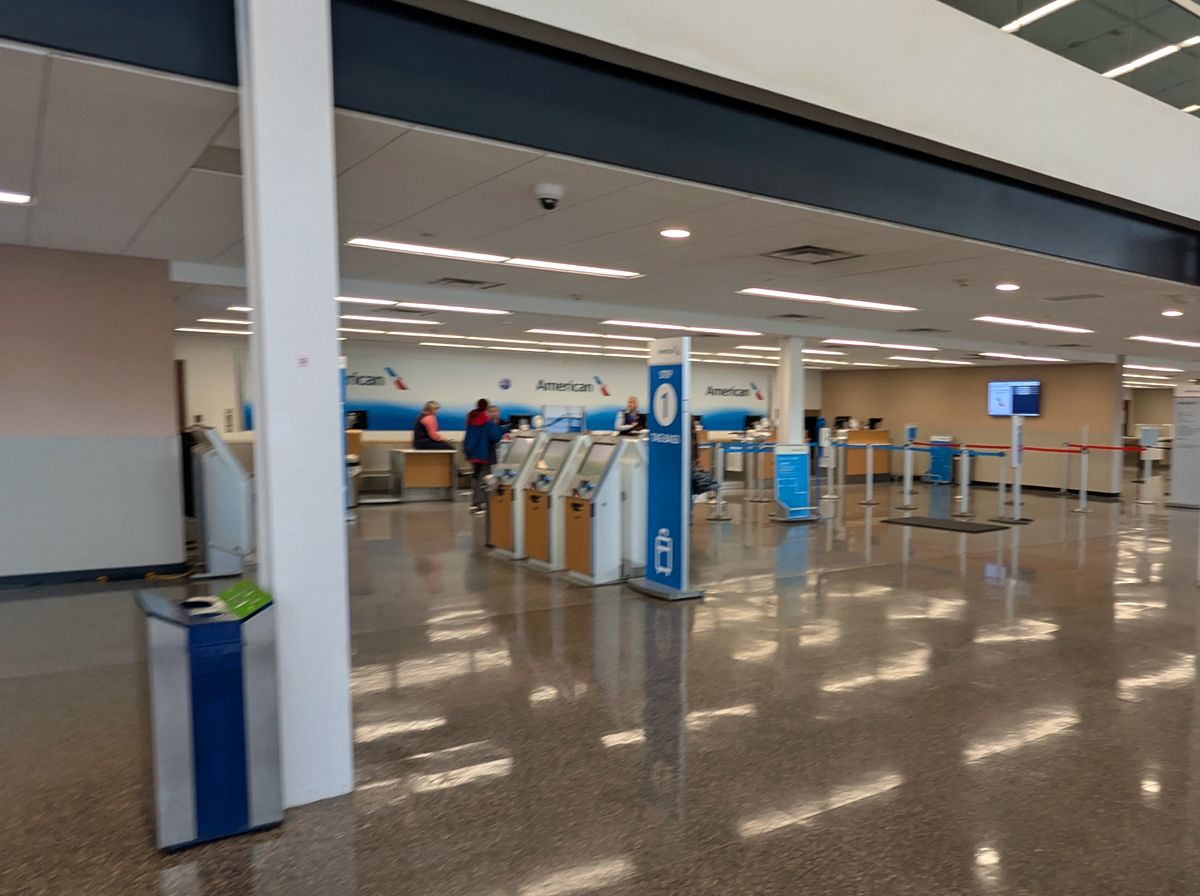 Three airline check-in desks under lit CHECK-IN signs, queue belts across a sunlit terrazzo floor and framed landscape photographs on the wall