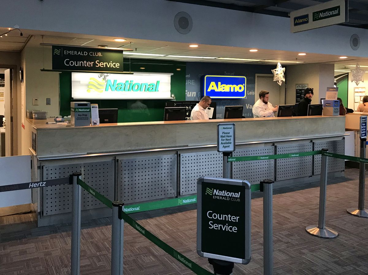 Three rental car counters with warm wood tops and slate blue panels in low evening light, tagged keys resting on the nearest counter