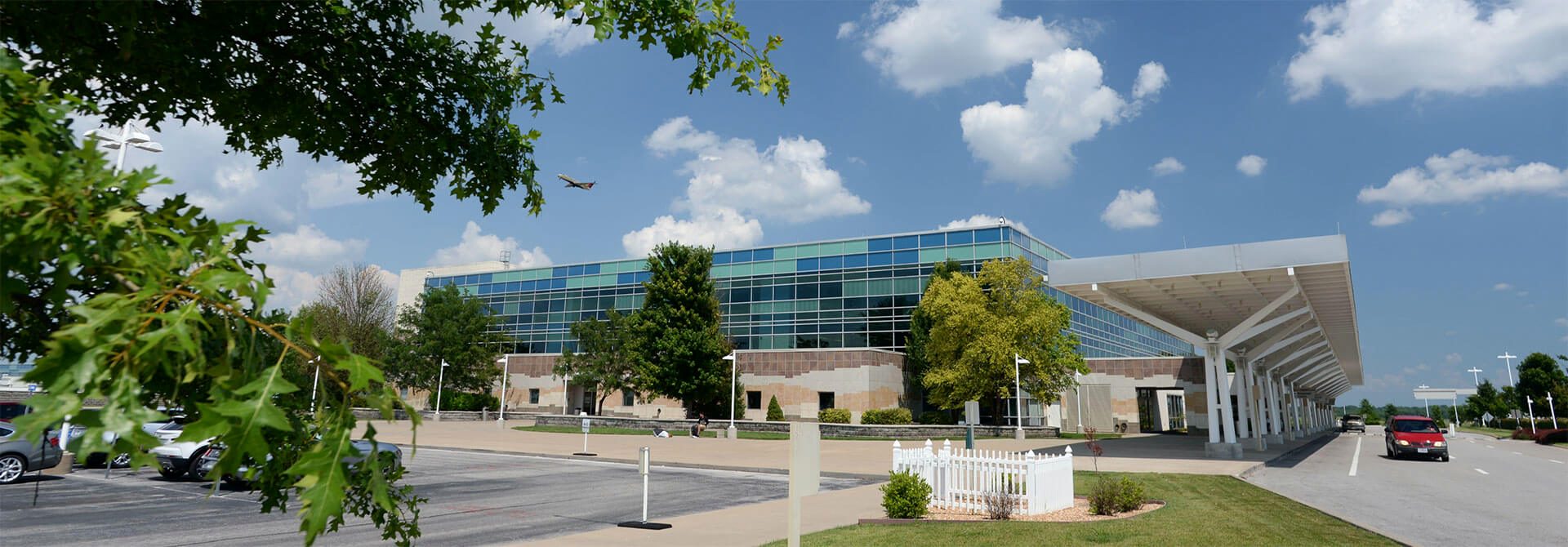 The Springfield-Branson National Airport terminal on a bright summer day, its curved glass facade framed by oak leaves, the covered drop-off canopy reaching toward the drive, and a departing aircraft climbing into a blue sky dotted with clouds